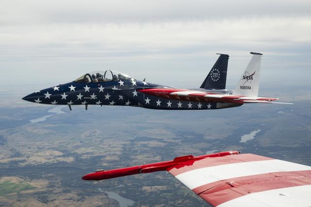 A red, white, and blue NASA F‑15 is shown beside another jet aircraft during a cross‑country flight. The full F‑15 is visible, while only a portion of the F/A‑18’s red‑and‑white wing appears in the frame.