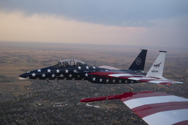 A red, white, and blue NASA F‑15 is shown beside another jet aircraft during a cross‑country flight. The full F‑15 is visible, while only a portion of the F/A‑18’s red‑and‑white wing appears in the frame.