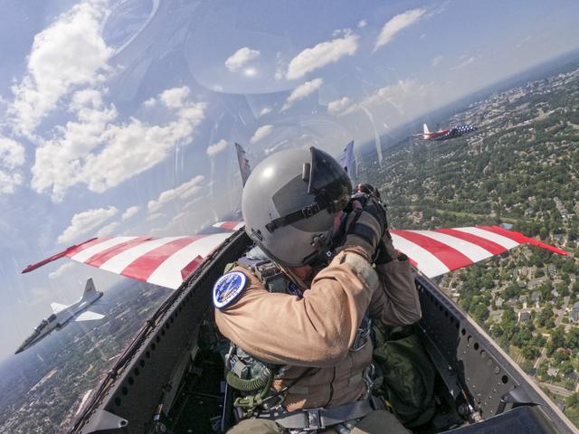 A man in a tan flight suit, flight gear, and helmet looks outside of the jet aircraft cockpit windows with a camera. Two other jet aircraft are seen outside of the cockpit window.