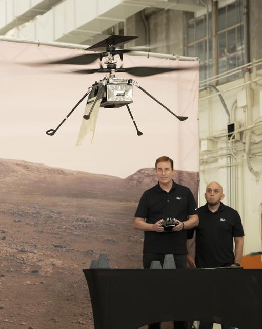 A small experimental helicopter hovers indoors in front of a backdrop showing a rocky, Mars‑like landscape. Two people wearing black shirts stand behind a table, watching the aircraft, with one holding a controller.