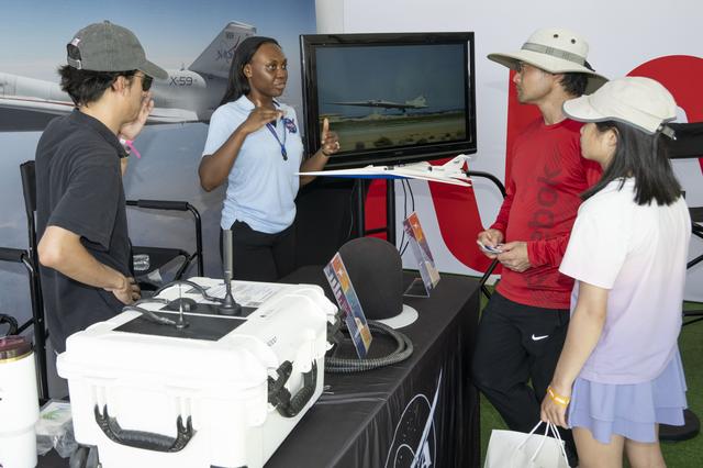 A NASA engineer speaks with visitors inside an exhibit booth, standing near a display screen showing an aircraft and a model of the X‑59 on the table.  