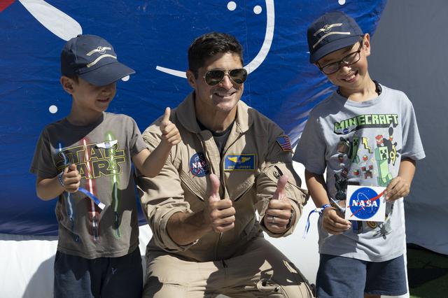 A NASA pilot in a tan flight suit kneels and smiles between two young children, all giving thumbs‑up, in front of a blue NASA backdrop at an outdoor event.