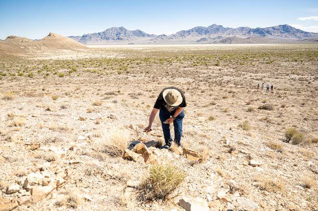 A man in a wide brimmed hat hits a rock with a hammer up on a hill. Down the hill in the background, a number of researchers stand looking out towards the mountains far off in the background. The ground is sparsely covered with brown and green shrubbery and off in the distance are mountains cutting up into a pale blue sky. 