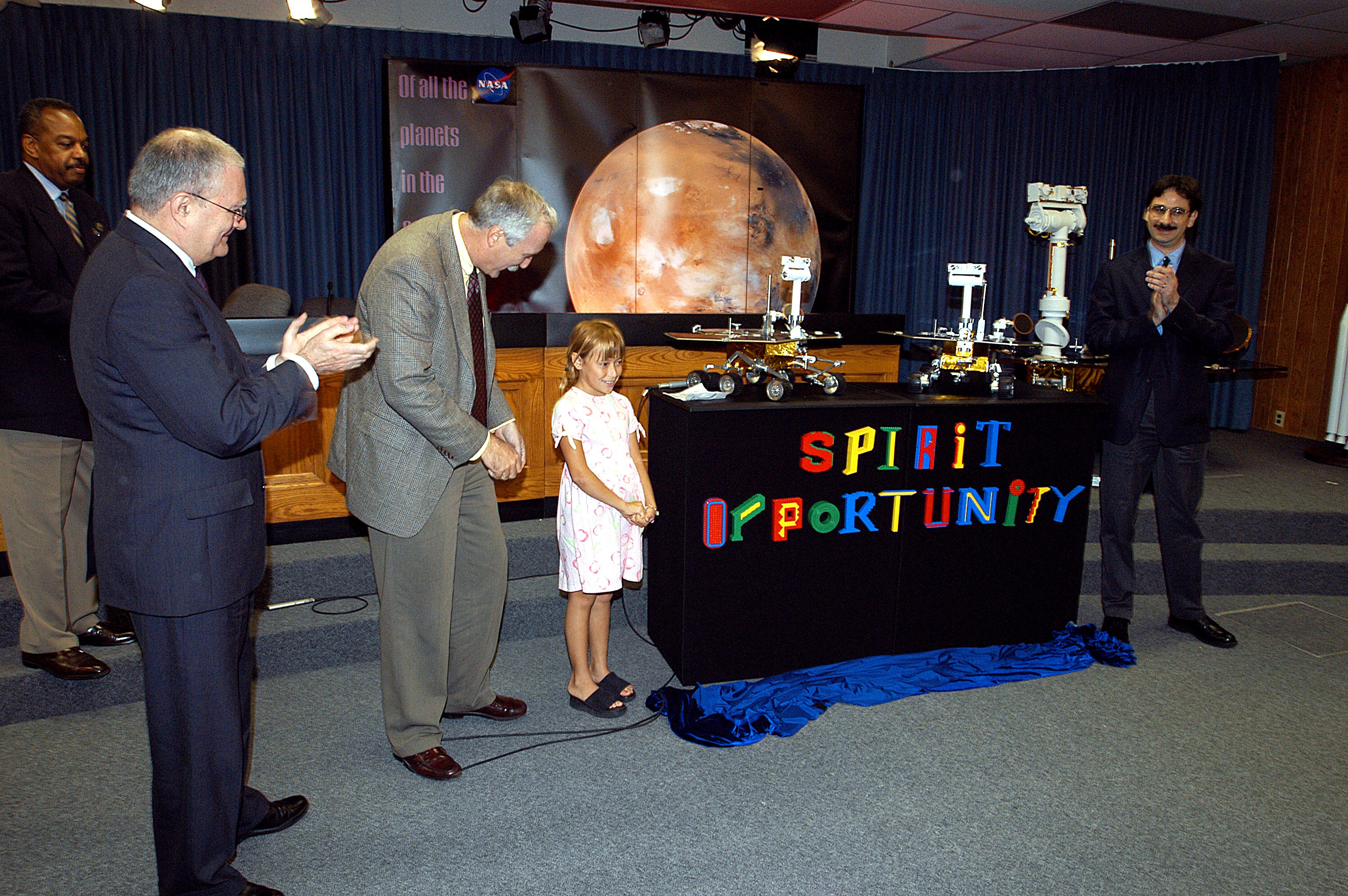 KENNEDY SPACE CENTER, FLA. - Nine-year-old Sofi Collis unveils the names of the Mars Exploration Rovers -- "Spirit" and "Opportunity" -- during a press conference. Participating in the press conference are, from left, Dr. John Marburger, science advisor to the President and director of the Office of Science and Technology Policy; NASA Administrator Sean O'Keefe; Sofi Collis, a third grade student from Arizona; and Brad Justus, LEGO Co. senior vice president. The names Sofi suggested were selected from more than 10,000 student entries in an essay contest managed for NASA by the LEGO Company. NASA's twin Mars Exploration Rovers are designed to study the history of water on Mars. These robotic geologists are equipped with a robotic arm, a drilling tool, three spectrometers, and four pairs of cameras that allow them to have a human-like, 3D view of the terrain. Each rover could travel as far as 100 meters in one day to act as Mars scientists' eyes and hands, exploring an environment where humans are not yet able to go. MER-A, with the rover Spirit aboard, is scheduled to launch on June 8 at 2:06 p.m. EDT, with two launch opportunities each day during a launch period that closes on June 24.