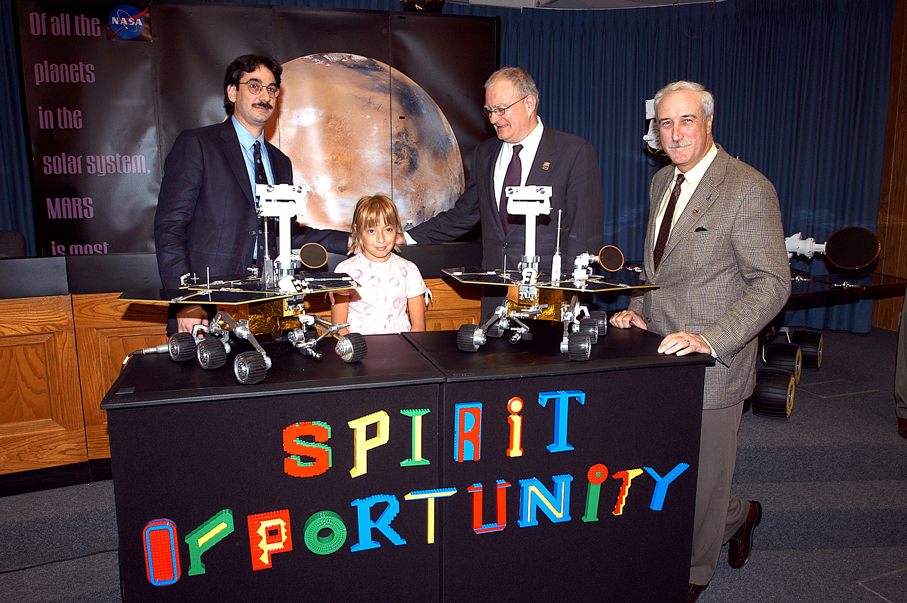 KENNEDY SPACE CENTER, FLA. - Nine-year-old Sofi Collis poses proudly with a banner displaying the names she selected for the Mars Exploration Rovers -- "Spirit" and "Opportunity" -- during a press conference. Participating in the press conference are, from left, Brad Justus, LEGO Co. senior vice president; Sofi Collis, a third grade student from Arizona; Dr. John Marburger, science advisor to the President and director of the Office of Science and Technology Policy; and NASA Administrator Sean O'Keefe. The names Sofi suggested were selected from more than 10,000 student entries in an essay contest managed for NASA by the LEGO Company. NASA's twin Mars Exploration Rovers are designed to study the history of water on Mars. These robotic geologists are equipped with a robotic arm, a drilling tool, three spectrometers, and four pairs of cameras that allow them to have a human-like, 3D view of the terrain. Each rover could travel as far as 100 meters in one day to act as Mars scientists' eyes and hands, exploring an environment where humans are not yet able to go. MER-A, with the rover Spirit aboard, is scheduled to launch on June 8 at 2:06 p.m. EDT, with two launch opportunities each day during a launch period that closes on June 24.