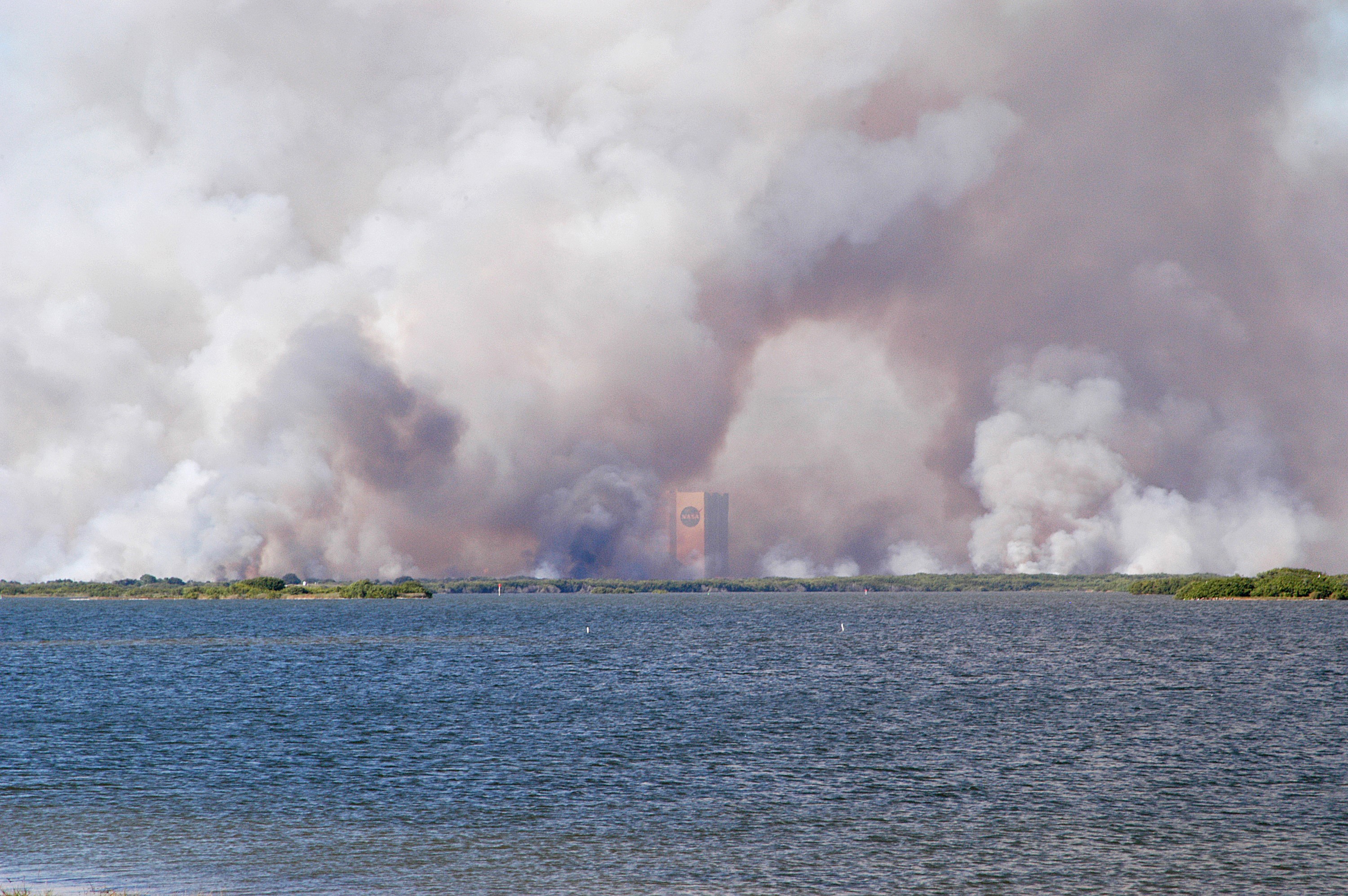 KENNEDY SPACE CENTER, FLA. - Smoke from a successful controlled burn near KSC’s Launch Complex 39 surrounds the Vehicle Assembly Building and spreads across the horizon. The water in the foreground is the Banana River.