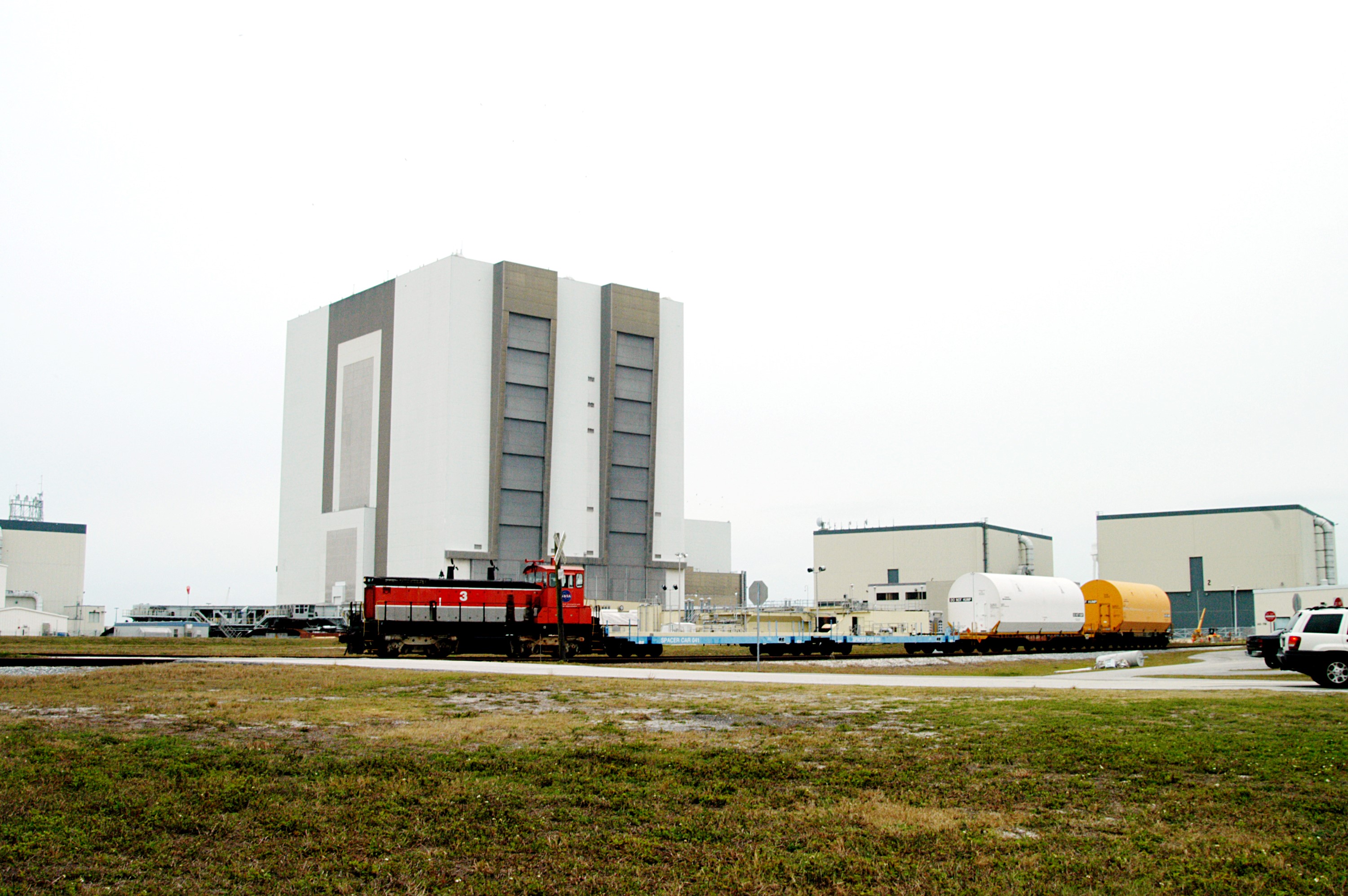 KENNEDY SPACE CENTER, FLA. - Moving past the 525-foot-high Vehicle Assembly Building, the red NASA engine pulls several containers enclosing segments of a solid rocket booster being returned to Utah for testing. The segments were part of the STS-114 stack. It is the first time actual flight segments that had been stacked for flight in the VAB are being returned for testing. They will undergo firing, which will enable inspectors to check the viability of the solid and verify the life expectancy for stacked segments.