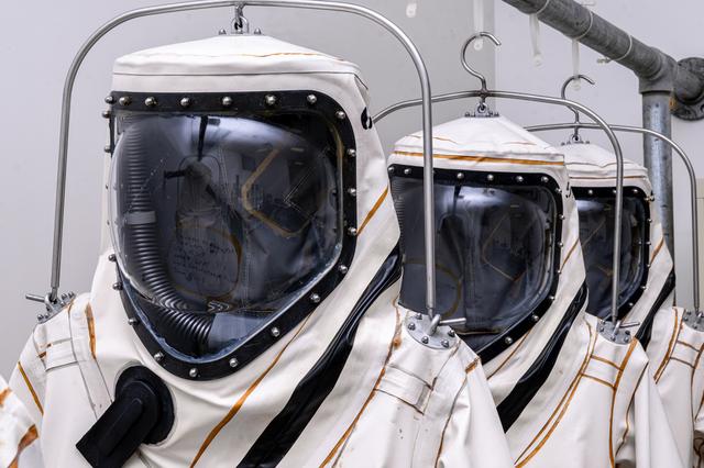 White and black protective suits hang on a black rack before technicians suit up to load propellant into a spacecraft. 