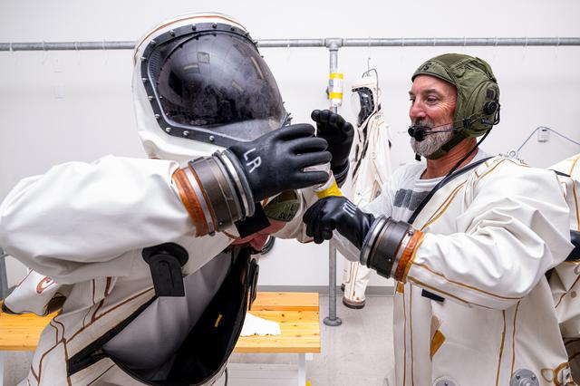 Two people wearing white protective suits and helmets in a preparation room; one adjusts the other’s gloved hand while benches and hanging equipment appear in the background.