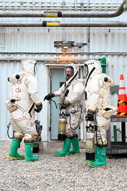 Three people stand outside a white building wearing white and black protective suits with green shoes.