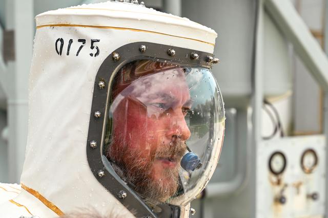 Close-up of a person in a white protective suit and clear face shield labeled “0175,” shown indoors beside gray metal equipment and cabling.