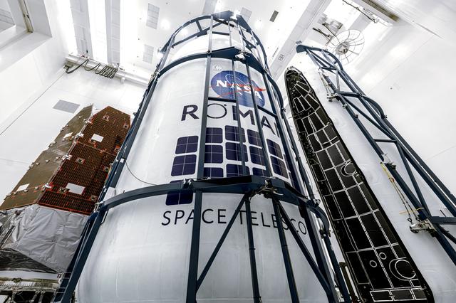 Angled view of the Roman Space Telescope wrapped in white covering with its mission logo visible, surrounded by tall blue support structures in the cleanroom.