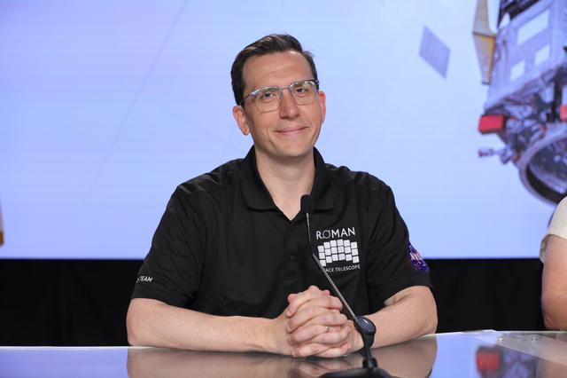A panelist sits behind a long, illuminated desk in front a video wall featuring NASA's Nancy Grace Roman Space Telescope during a NASA news conference. 