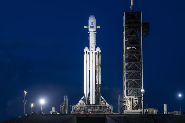 A white SpaceX Falcon Heavy rocket stands vertical on the black launch pad with a predawn sky in the background.