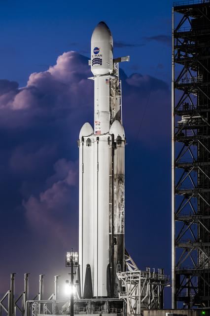 A white SpaceX Falcon Heavy rocket stands vertical on the black launch pad with a predawn sky in the background.