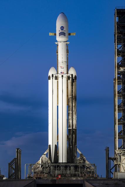 A white SpaceX Falcon Heavy rocket stands vertical on the black launch pad with a predawn sky in the background.