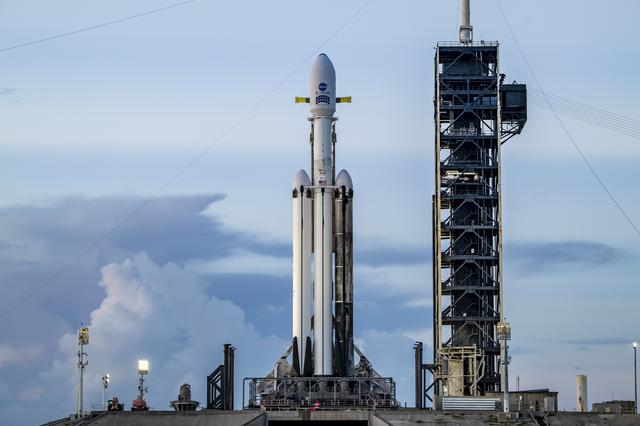 A white SpaceX Falcon Heavy rocket stands vertical on the black launch pad with a predawn sky in the background.