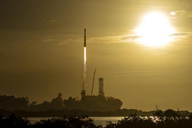 A rocket lifts off from a coastal launch pad, sending bright flames and a plume of white smoke across the ground.