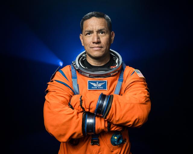 NASA astronaut Frank Rubio is pictured wearing an orange Orion Crew Survival System suit in a close-up shot. He is standing in front of a dark backdrop, illuminated with blue lights, with his arms crossed in front of his chest.