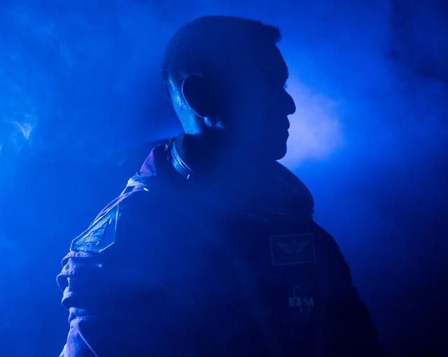 A side-profile, close-up silhouette of NASA astronaut Frank Rubio is pictured with blue lights and fog illuminating the background. He is pictured in an Orion Crew Survival System suit without the helmet.