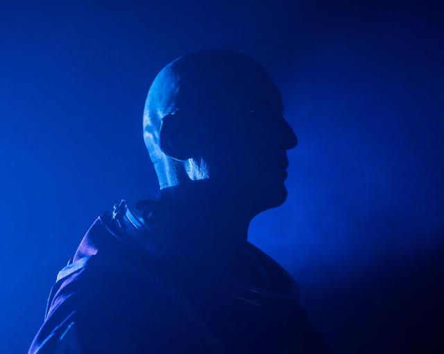 A side-profile, close-up silhouette of ESA (European Space Agency) astronaut Luca Parmitano is pictured with blue lights and fog illuminating the background. He is pictured in an Orion Crew Survival System suit without the helmet.
