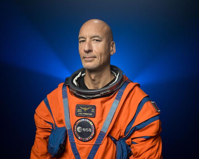 ESA (European Space Agency) astronaut Luca Parmitano is pictured wearing an orange Orion Crew Survival System suit in a close-up shot. He is standing in front of a dark backdrop, illuminated with blue lights, with his arms at his side.