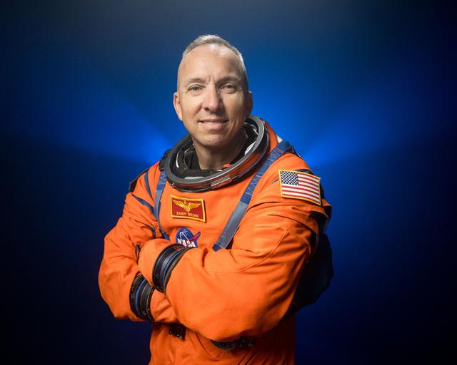 NASA astronaut Randy Bresnik is pictured wearing an orange Orion Crew Survival System suit in a close-up shot. He is standing in front of a dark backdrop, illuminated with blue lights, with his arms crossed in front of his chest.