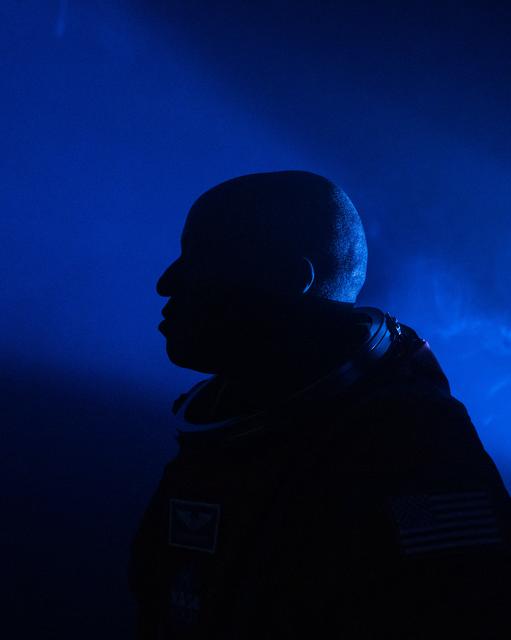 A side-profile, close-up silhouette of NASA astronaut Andre Douglas is pictured with blue lights and fog illuminating the background. He is pictured in an Orion Crew Survival System suit without the helmet.