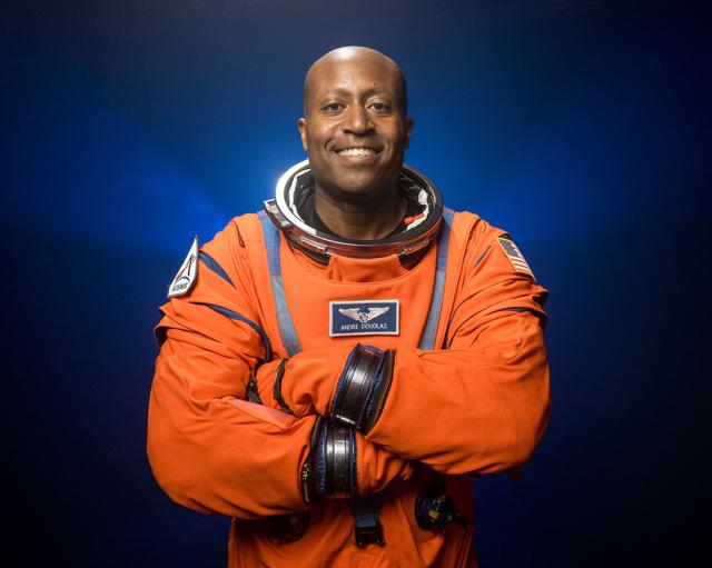 NASA astronaut Andre Douglas is pictured wearing an orange Orion Crew Survival System suit in a close-up shot. He is standing in front of a dark backdrop, illuminated with blue lights, with his arms crossed in front of his chest.