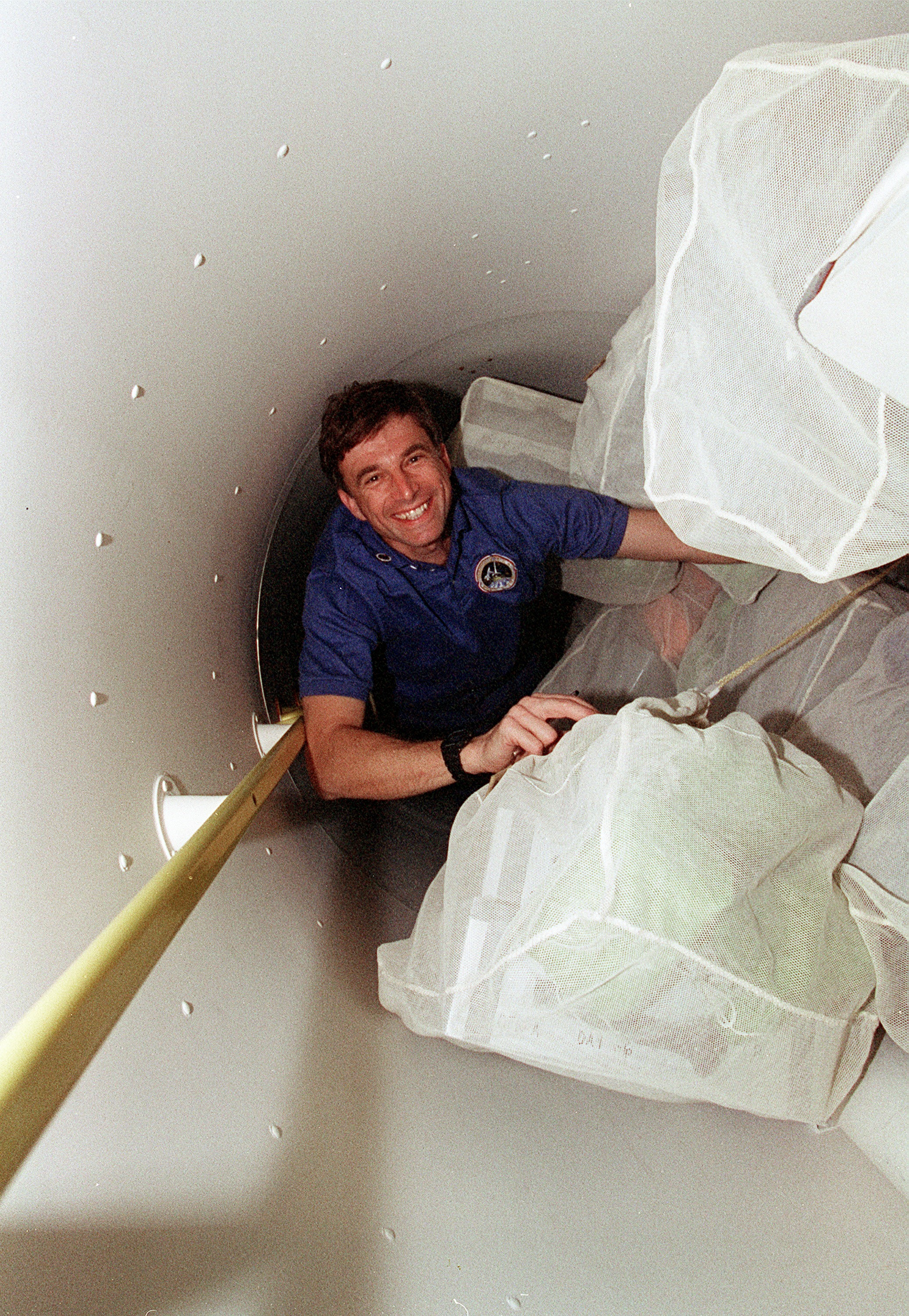 Astronauts move through narrow space in tunnel adapter filled with transfer bag