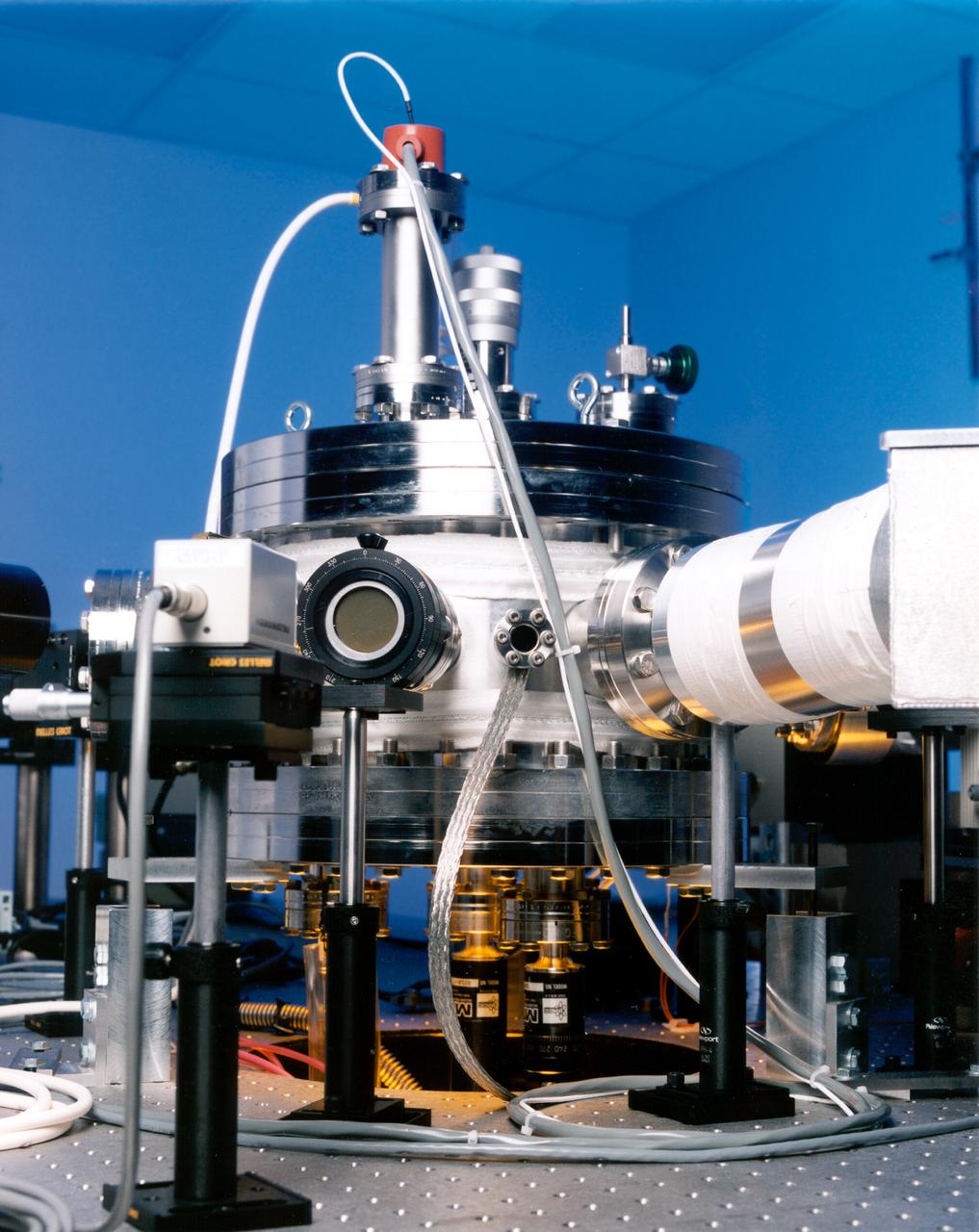 Optical prots ring the Electrostatic Levitator (ESL) vacuum chamber to admit light from the heating laser (the beam passes through the window at left), poisitioning lasers (one port is at center), and lamps (such as the deuterium arc lamp at right), and to allow diagnostic instruments to view the sample. The ESL uses static electricity to suspend an object (about 2-3 mm in diameter) inside a vacuum chamber while a laser heats the sample until it melts. This lets scientists record a wide range of physical properties without the sample contacting the container or any instruments, conditions that would alter the readings. The Electrostatic Levitator is one of several tools used in NASA's microgravity materials science program. 