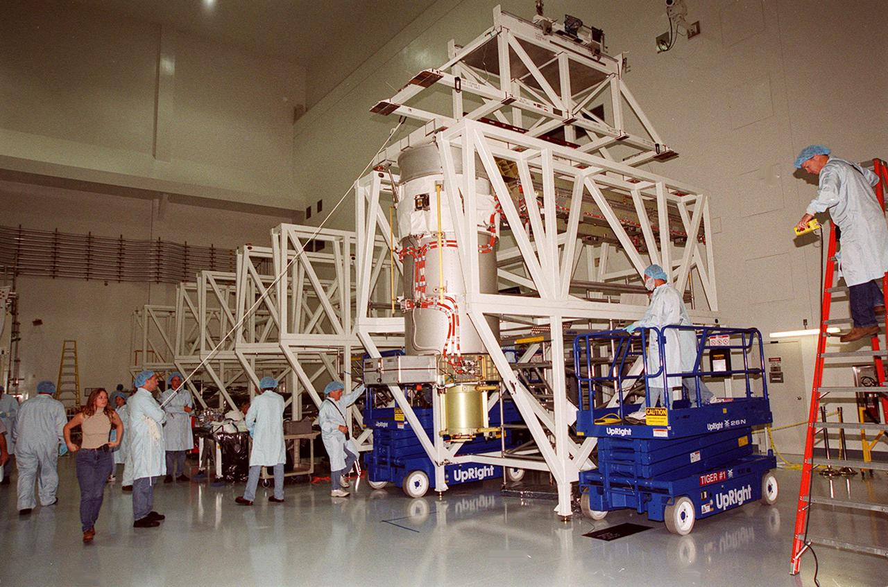 Workers in the Space Station Processing Facility get ready to attach an overhead crane (center top) to the solar array below it to move the array for installation onto the Integrated Equipment Assembly (IEA). A component of the International Space Station, the solar array is the second one being installed on the IEA. The arrays are scheduled to be launched on mission STS-97 in late November along with the P6 truss. The Station’s electrical power system (EPS) will use eight photovoltaic solar arrays to convert sunlight to electricity. Each of the eight solar arrays will be 112 feet long by 39 feet wide. The solar arrays are mounted on a “blanket” that can be folded like an accordion for delivery. Once in orbit, astronauts will deploy the blankets to their full size. Gimbals will be used to rotate the arrays so that they will face the Sun to provide maximum power to the Space Station