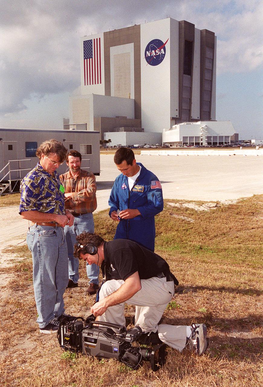 Across from the Vehicle Assembly Building and Launch Control Center, Steve Thomas (left), host of This Old House, and Norm Abram (second from left), master carpenter on the series, watch as a a videographer (in front) checks his camera. With them is astronaut John Herrington. The cast and crew of This Old House are filming at KSC for an episode of the show. Herrington is accompanying the film crew on their tour of KSC