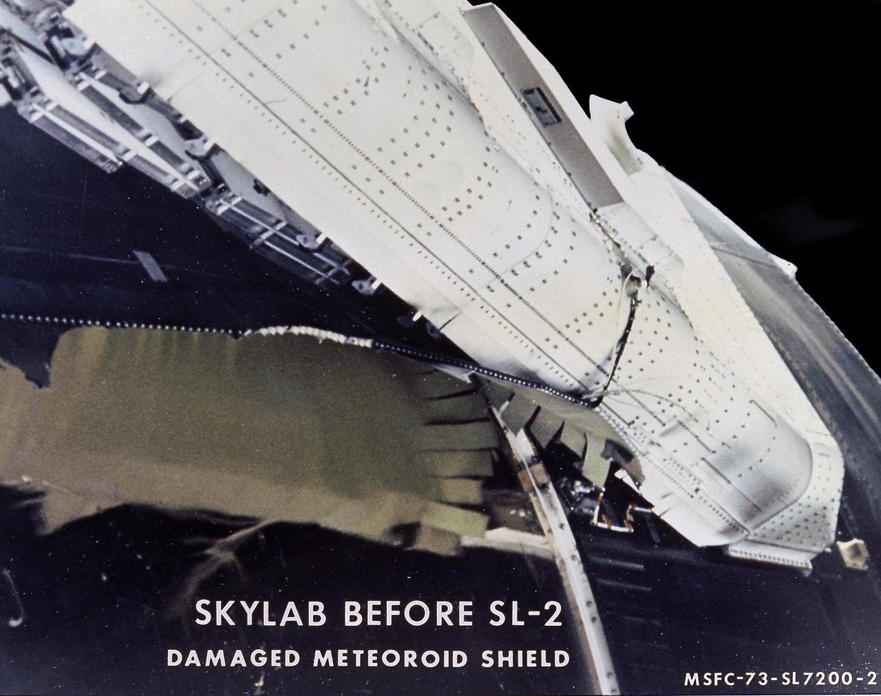 The Saturn V vehicle, carrying the unmarned orbital workshop for the Skylab-1 mission, lifted off successfully and all systems performed normally. Sixty-three seconds into flight, engineers in the operation support and control center saw an unexpected telemetry indication that signalled that damages occurred on one solar array and the micrometeoroid shield during the launch. The micrometeoroid shield, a thin protective cylinder surrounding the workshop protecting it from tiny space particles and the sun's scorching heat, ripped loose from its position around the workshop. This caused the loss of one solar wing and jammed the other. Still unoccupied, the Skylab was stricken with the loss of the heat shield and sunlight beat mercilessly on the lab's sensitive skin. Internal temperatures soared, rendering the station uninhabitable, threatening foods, medicines, films, and experiments. This image, taken during a fly-around inspection by the Skylab-2 crew, shows the damaged meteoroid shield being held by a thin aluminum strap entangled with green-hued remnants of the lost heat shield. The Marshall Space Flight Center (MSFC) developed, tested, rehearsed, and approved three repair options. These options included a parasol sunshade and a twin-pole sunshade to restore the temperature inside the workshop, and a set of metal cutting tools to free the jammed solar panel.