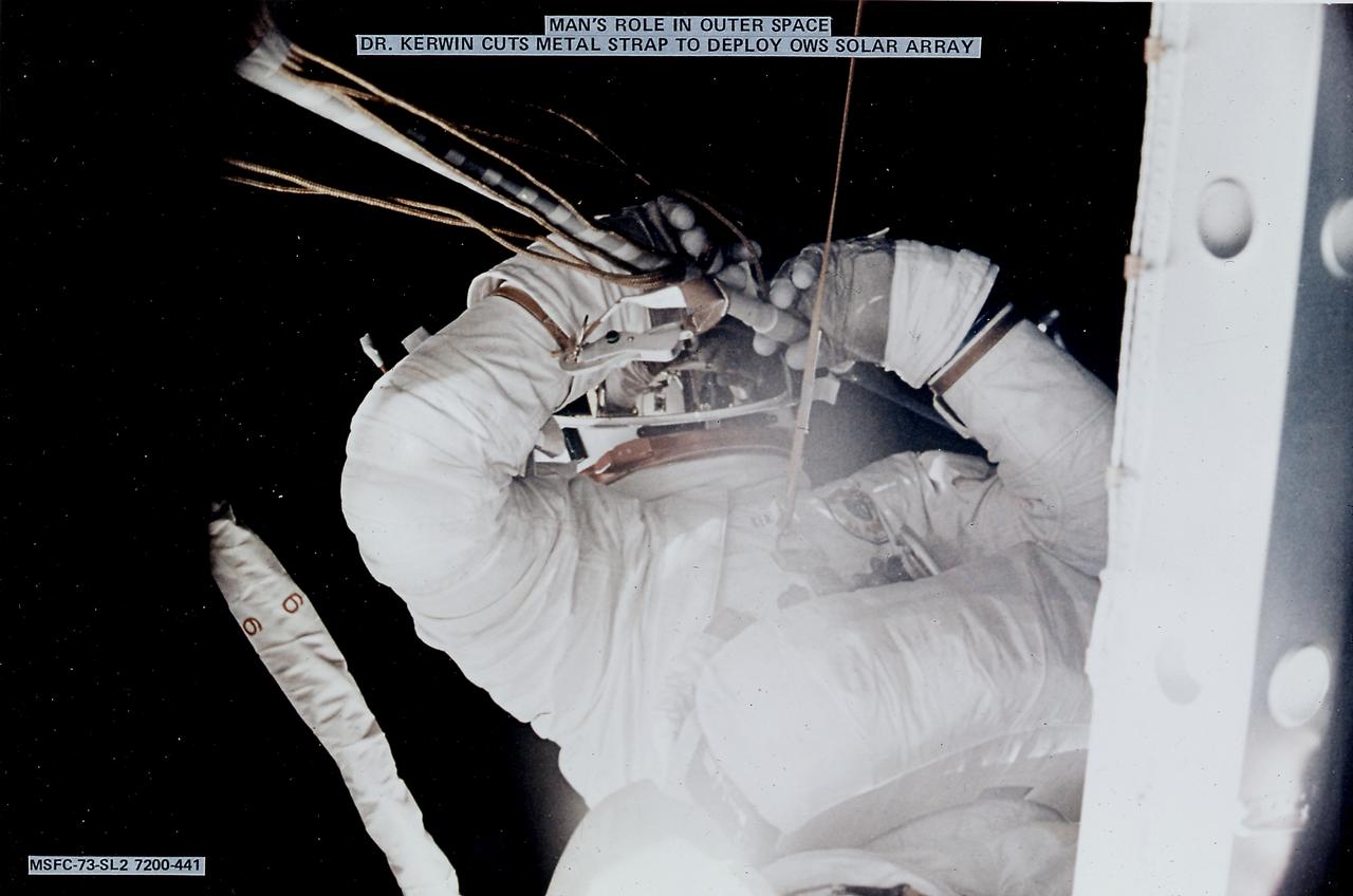The Saturn V vehicle, carrying the unmarned orbital workshop for the Skylab-1 mission, lifted off successfully and all systems performed normally. Sixty-three seconds into the flight, engineers in the operation support and control center saw an unexpected telemetry indication that signalled that damages occurred on one solar array and the micrometeoroid shield during the launch. The micrometeoroid shield, a thin protective cylinder surrounding the workshop protecting it from tiny space particles and the sun's scorching heat, ripped loose from its position around the workshop. This caused the loss of one solar wing and jammed the other. Still unoccupied, the Skylab was stricken with the loss of the heat shield and sunlight beat mercilessly on the lab's sensitive skin. Intrnal temperatures soared, rendering the station uninhabitable, threatening foods, medicines, films, and experiments. This image shows astronaut Kerwin cutting the metal strap to free and deploy the Orbital Workshop solar array. Kerwin used special cutting tools developed by engineers at the Marshall Space Flight Center (MSFC). The MSFC had a major role in developing the procedures to repair the damaged Skylab. 