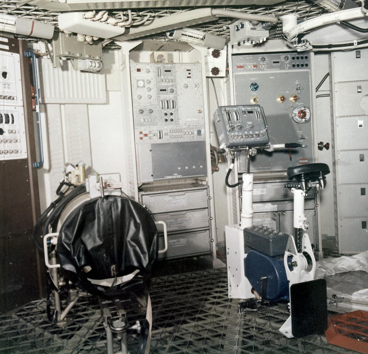 The Orbital Workshop internal arrangement shown here is the medical experimental equipment. In this view, looking from the wardroom area, are the lower-body Negative Pressure (Experiment M092) unit, left, and the ergometer for the vectorcardiograph (Experiment - M093). Both are used in several ways to keep check on the astronauts' condition and tolerance in extended weightlessness. The 1-G trainer permits the astronauts to get experience with all of the equipment and operations except the absence of gravity. 