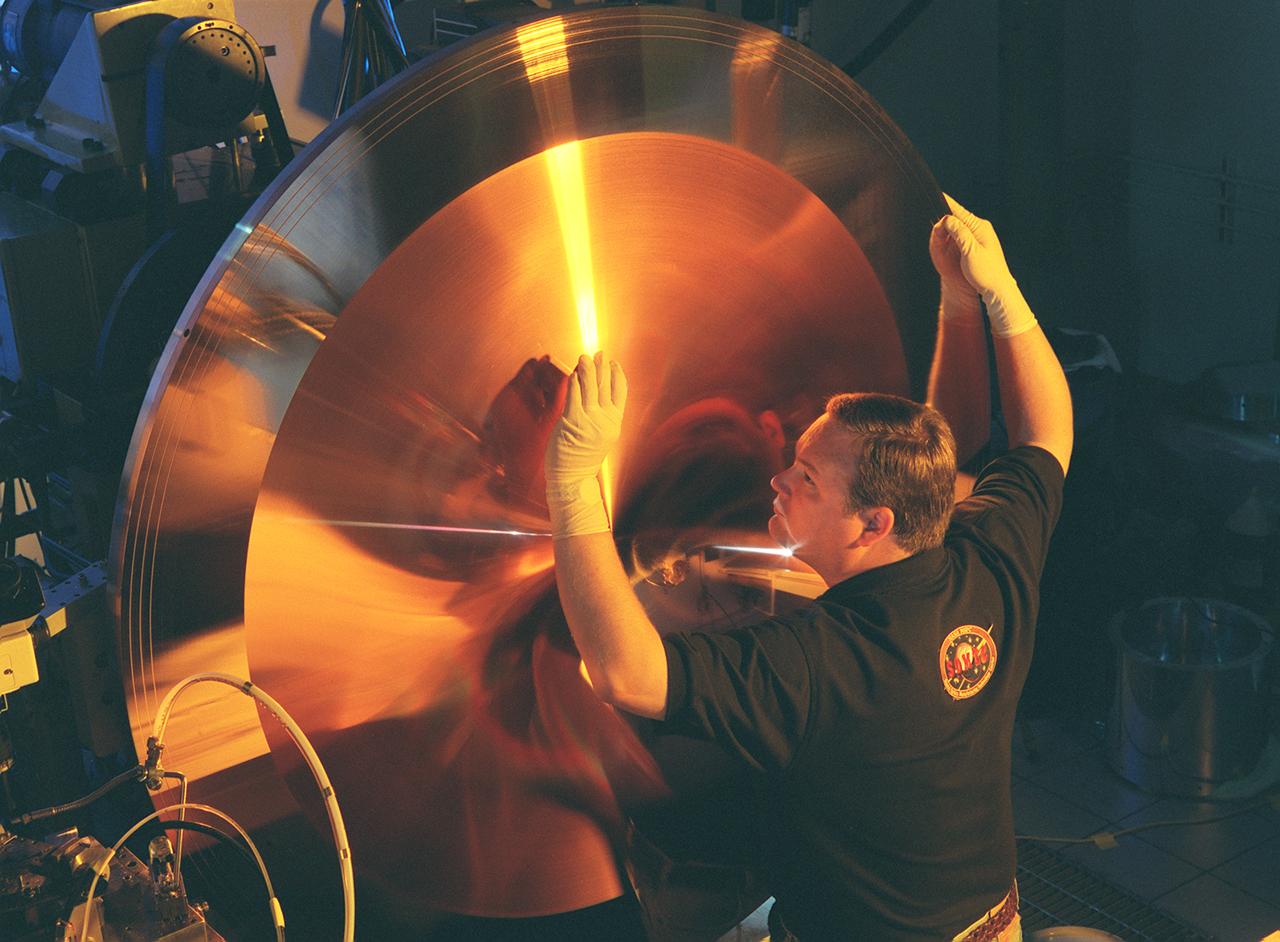 This photograph shows Wes Brown, Marshall Space Flight Center's (MSFC's) lead diamond tuner, an expert in the science of using diamond-tipped tools to cut metal, inspecting the mold's physical characteristics to ensure the uniformity of its more than 6,000 grooves. This king-size copper disk, manufactured at the Space Optics Manufacturing and Technology Center (SOMTC) at MSFC, is a special mold for making high resolution monitor screens. This master mold will be used to make several other molds, each capable of forming hundreds of screens that have a type of lens called a fresnel lens. Weighing much less than conventional optics, fresnel lenses have multiple concentric grooves, each formed to a precise angle, that together create the curvature needed to focus and project images. The MSFC leads NASA's space optics manufacturing technology development as a technology leader for diamond turning. The machine used to manufacture this mold is among many one-of-a-kind pieces of equipment of MSFC's SOMTC.