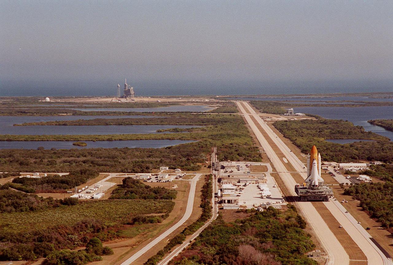 KENNEDY SPACE CENTER, FLA. -- Space Shuttle <a href='.._.._subjects_atlantis.htm'> Atlantis<_a> (right) inches its way at 1 mph atop the crawler-transporter back to the <a href='.._.._subjects_vab.htm'> Vehicle Assembly Building<_a> from <a href='.._.._subjects_lc39a.htm'> Launch Pad 39A<_a> (upper left). A panorama view from the top of the VAB shows the proximity of the pad to the Atlantic Ocean (background) plus the 3.4-mile crawlerway leading from the pad to the VAB. The water areas on both sides of the crawlerway are part of the Banana River. In the VAB workers will conduct inspections, make continuity checks and conduct X-ray analysis on the 36 solid rocket booster cables located inside each booster’s external system tunnel. An extensive evaluation of NASA’s SRB cable inventory revealed conductor damage in four (of about 200) cables on the shelf. Shuttle managers decided to prove the integrity of the system tunnel cables already on Atlantis before launching. The launch has been rescheduled no earlier than Feb. 6