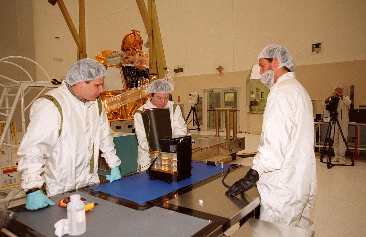 In the Spacecraft Assembly and Encapsulation Facility 2 (SAEF 2), workers check the Thermal Emission Imaging System (THEMIS) before attaching to the 2001 Mars Odyssey Orbiter (background). THEMIS will map the mineralogy and morphology of the Martian surface using a high-resolution camera and a thermal infrared imaging spectrometer. The orbiter will carry three science instruments: THEMIS, the Gamma Ray Spectrometer (GRS), and the Mars Radiation Environment Experiment (MARIE). The GRS will achieve global mapping of the elemental composition of the surface and determine the abundance of hydrogen in the shallow subsurface. The MARIE will characterize aspects of the near-space radiation environment with regards to the radiation-related risk to human explorers. The Mars Odyssey Orbiter is scheduled for launch on April 7, 2001, aboard a Delta 7925 rocket from Launch Pad 17-A, Cape Canaveral Air Force Station