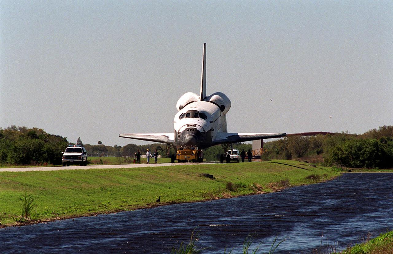 Accompanied by workers and Security, Atlantis moves along the tow-way from the KSC Shuttle Landing Facility to the Orbiter Processing Facility. Atlantis returned from California atop a Shuttle Carrier Aircraft after its Feb. 19 landing at Edwards Air Force Base, concluding mission STS-98. The ferry flight began March 1; unfavorable weather conditions kept it on the ground at Altus AFB, Okla., until it could return to Florida. In the Orbiter Processing Facility, Atlantis will be prepared for mission STS-104, the 10th construction flight to the International Space Station, scheduled to launch June 8