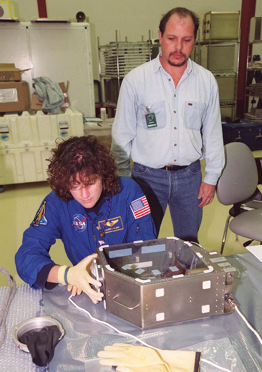 KENNEDY SPACE CENTER, Fla. -- Under the watchful eyes of a trainer at SPACEHAB, Cape Canaveral, Fla., STS-107 Mission Specialist Laurel Clark checks out a glove box experiment for the June 2002 mission. STS-107 has two payload elements, the Double Module in its first flight into space and a Hitchhiker payload. The experiments range from material sciences to life sciences (many rats). The Hitchhiker carrier system is modular and expandable in accordance with payload requirements. Hitchhiker experiments are housed in canisters or attached to mounting plates. The Hitchhiker canister comes in two varieties--the Hitchhiker Motorized Door Canister and the Sealed Canisters