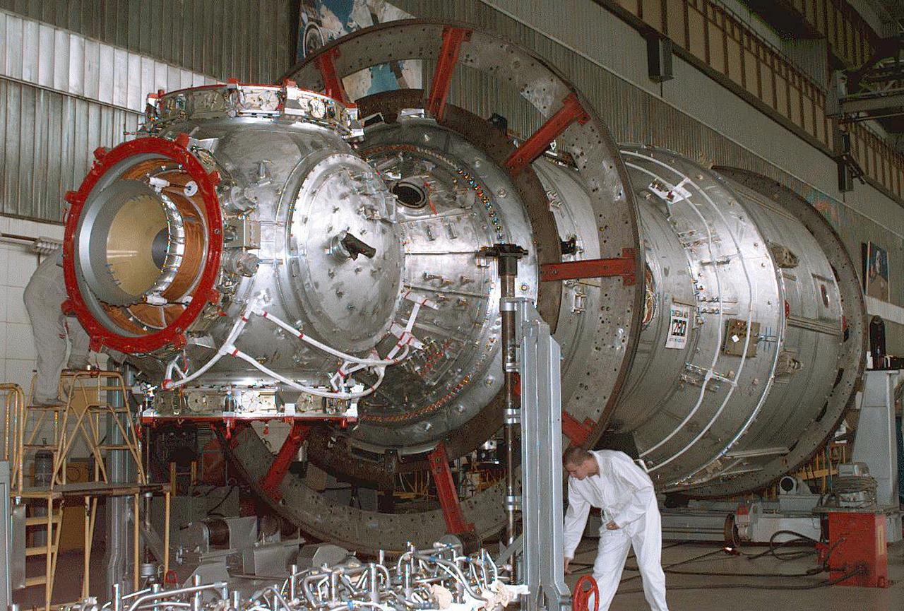 The Zvezda Service Module, the first Russian contribution and third element to the International Space Station (ISS), is shown under construction in the Krunichev State Research and Production Facility (KhSC) in Moscow. Russian technicians work on the module shortly after it completed a pressurization test. In the foreground is the forward portion of the module, including the spherical transfer compartment and its three docking ports. The forward port docked with the cornected Functional Cargo Block, followed by Node 1. Launched via a three-stage Proton rocket on July 12, 2000, the Zvezda Service Module serves as the cornerstone for early human habitation of the Station, providing living quarters, life support system, electrical power distribution, data processing system, flight control system, and propulsion system. It also provides a communications system that includes remote command capabilities from ground flight controllers. The 42,000-pound module measures 43 feet in length and has a wing span of 98 feet. Similar in layout to the core module of Russia's Mir space station, it contains 3 pressurized compartments and 13 windows that allow ultimate viewing of Earth and space.