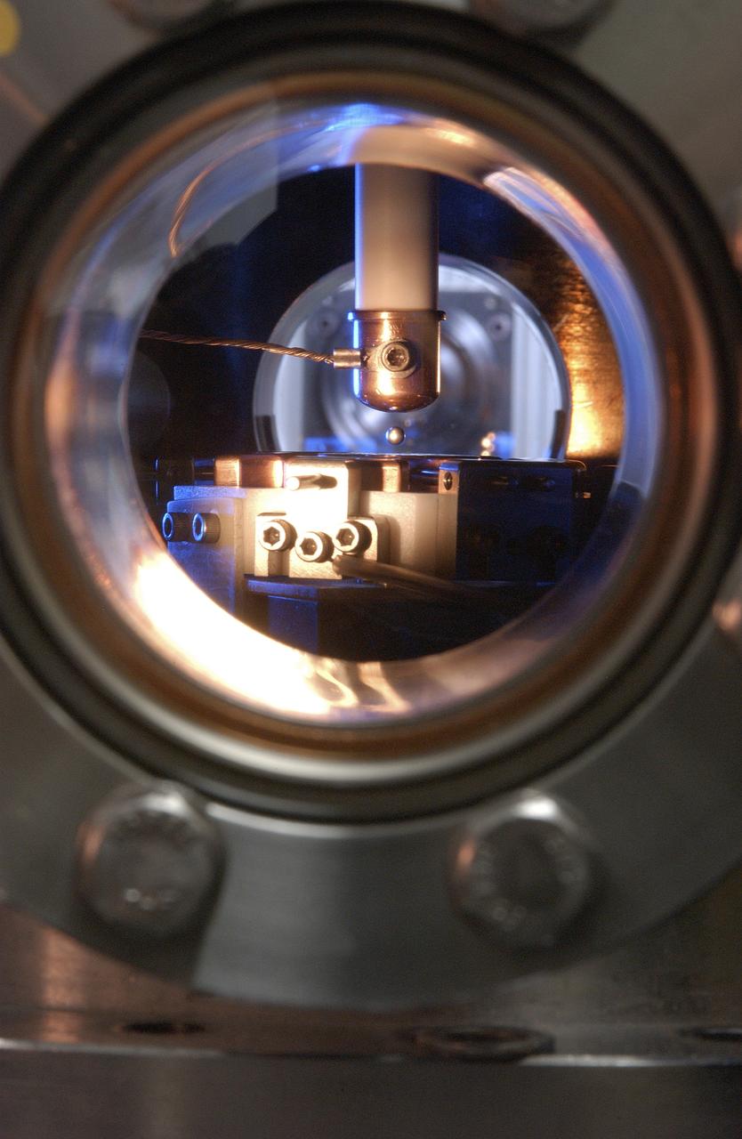 This Photo, which appeared on the July cover of `Physics Today', is of the Electrostatic Levitator (ESL) at NASA's Marshall Space Flight Center (MSFC). The ESL uses static electricity to suspend an object (about 3-4 mm in diameter) inside a vacuum chamber allowing scientists to record a wide range of physical properties without the sample contracting the container or any instruments, conditions that would alter the readings. Once inside the chamber, a laser heats the sample until it melts. The laser is then turned off and the sample cools, changing from a liquid drop to a solid sphere. In this particular shot, the ESL contains a solid metal sample of titanium-zirconium-nickel alloy. Since 1977, the ESL has been used at MSFC to study the characteristics of new metals, ceramics, and glass compounds. Materials created as a result of these tests include new optical materials, special metallic glasses, and spacecraft components.