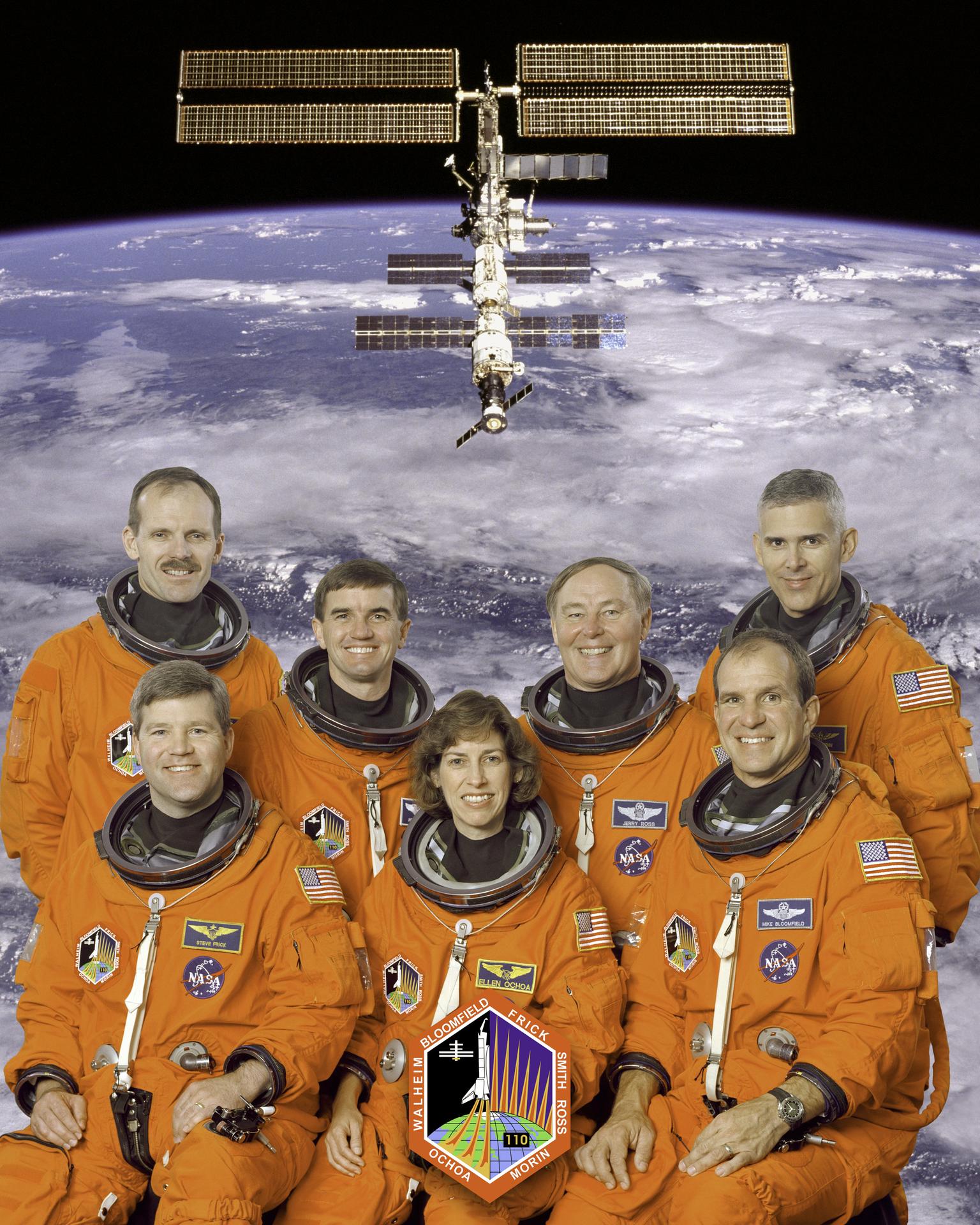 Seven astronauts pose for crew photo in orange spacesuits with earth and satellite background