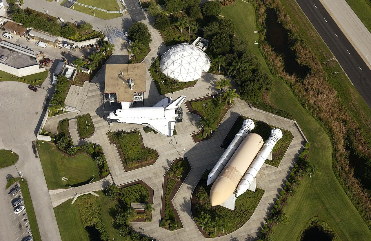 An aerial view of the KSC Visitor Complex shows the Shuttle Plaza, with the full-size replica of an orbiter.  Nearby is the display of solid rocket boosters and an external tank.                               