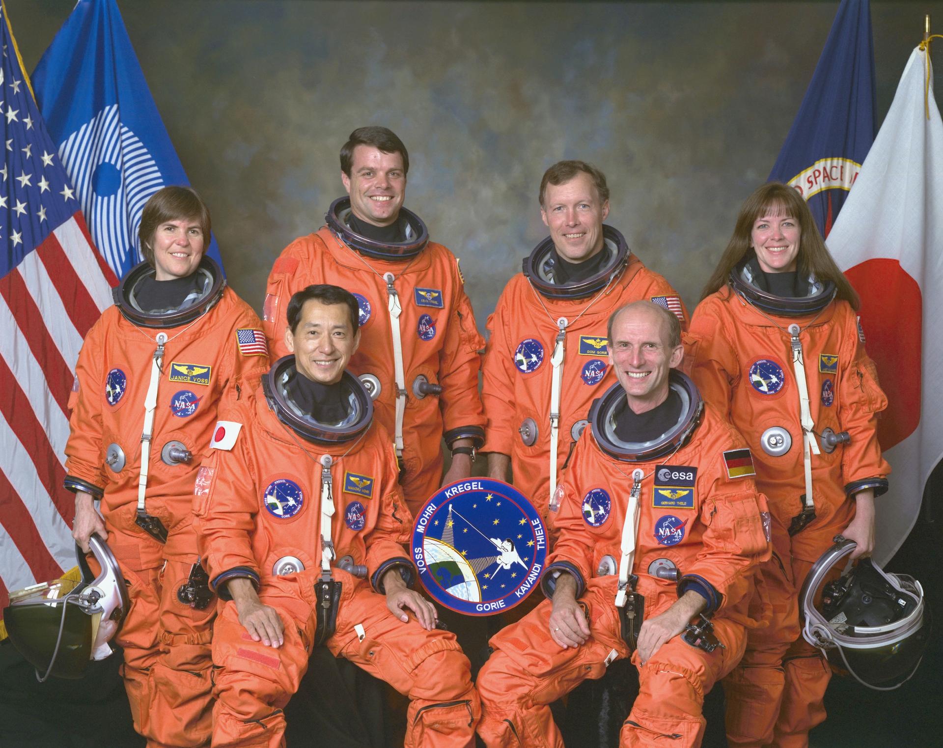 Six astronauts pose in orange spacesuits in front of flags