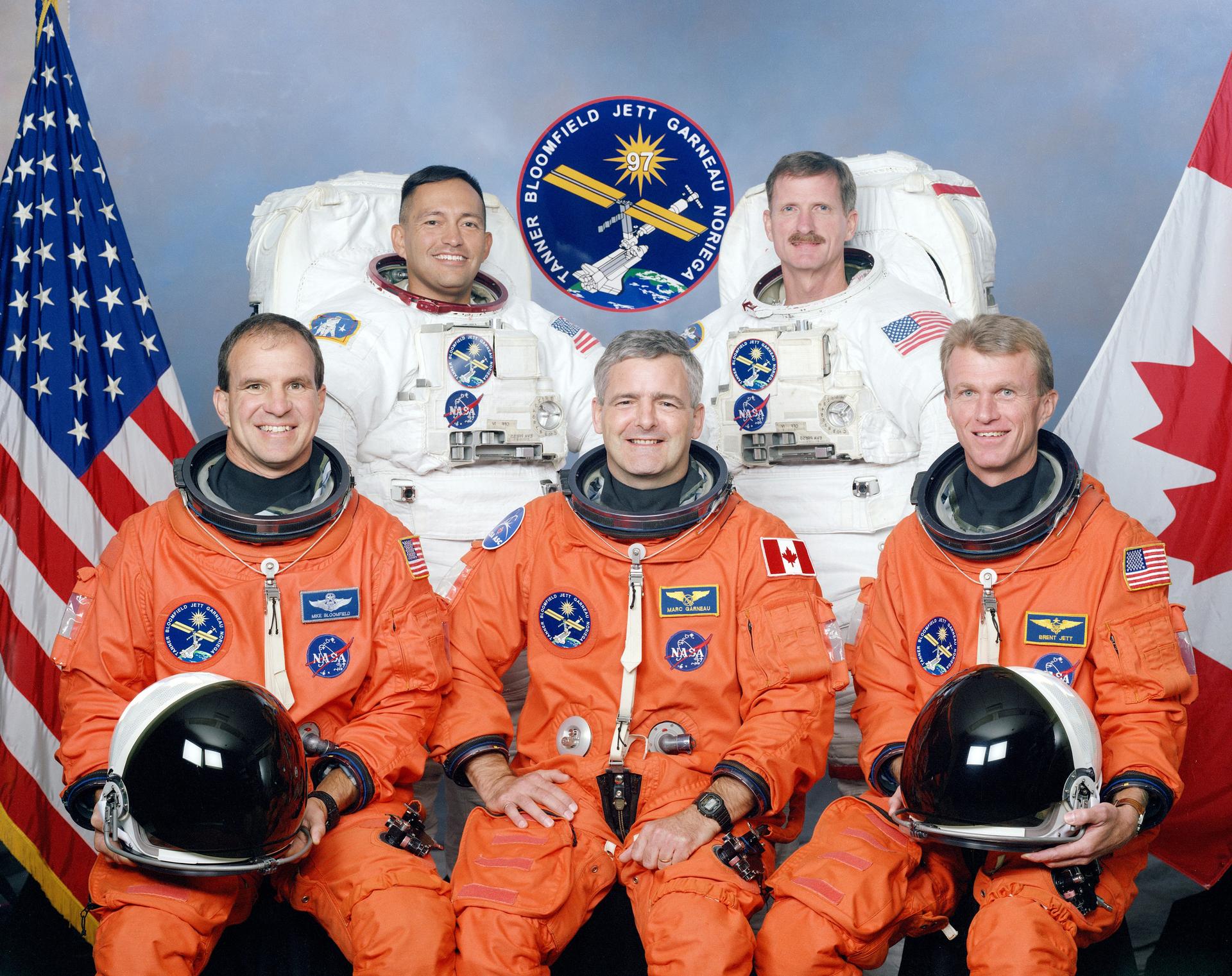 Five astronauts in orange and white spacesuits pose in front of US and Canadian flag.