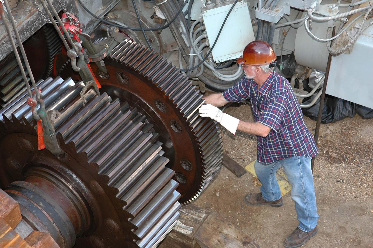 KENNEDY SPACE CENTER, FLA. -   A worker maneuvers the gear into position on the Crawler-Transporter.  At far left is the giant-sized sprocket.  The drive sprocket turns the belt on the CT.  The sprocket is mated to the gear that attaches to the drive motor.  The CT travels on eight tracked tread belts, each containing 57 tread belt shoes, for a combined weight of 957,600 pounds.  The CT carries the Space Shuttle atop its Mobile Launcher Platform, adding another 12 million pounds, from the Vehicle Assembly Building to the launch pad.  NASA and United Space Alliance (USA) CT system engineers and USA technicians are repairing the sprockets and rollers on each belt before new shoes are installed.  Replacement of the sprockets, gears and shoes is part of the most extensive maintenance work performed on the CT in its history.
