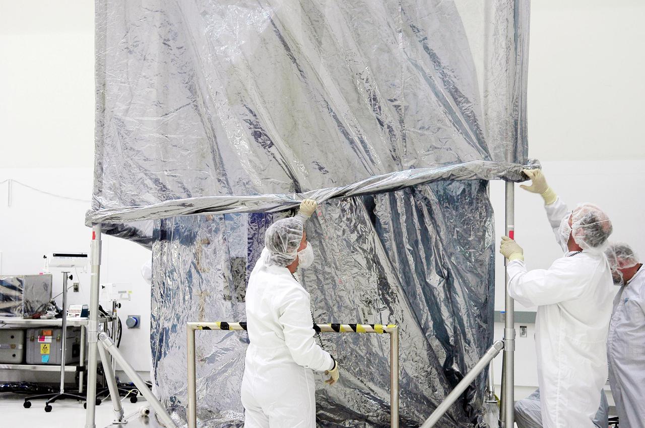 KENNEDY SPACE CENTER, FLA. -  At Astrotech Space Operations in Titusville, Fla., workers lower a second protective cover around the Deep Impact spacecraft.  Deep Impact is being prepared for a move to the Hazardous Fuel Building and eventually shipment to Launch Pad 17-B, Cape Canaveral Air Force Station. At Astrotech Space Operations in Titusville, Fla., workers help guide an overhead crane holding a protective cover that will be placed over the Deep Impact spacecraft below. Deep Impact is being prepared for a move to the Hazardous Fuel Building and eventually shipment to Launch Pad 17-B, Cape Canaveral Air Force Station. Launch of Deep Impact is scheduled no earlier than Jan. 12.  A NASA Discovery mission, Deep Impact will probe beneath the surface of Comet Tempel 1 on July 4, 2005, when the comet is 83 million miles from Earth, and reveal the secrets of its interior.   After releasing a 3- by 3-foot projectile to crash onto the surface, Deep Impact’s flyby spacecraft will collect pictures and data of how the  crater forms, measuring the crater’s depth and diameter, as well as the composition of the interior of the crater and any material thrown out, and determining the changes in natural outgassing produced by the impact.  It will send the data back to Earth through the antennas of the Deep Space Network.