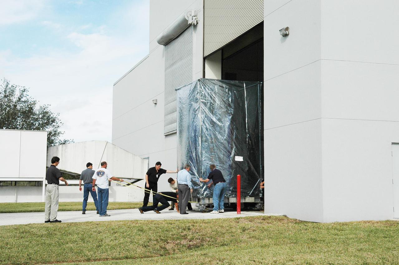 KENNEDY SPACE CENTER, FLA. -  At Astrotech Space Operations in Titusville, Fla., workers push and pull the moveable stand holding the Deep Impact spacecraft to get it through the door.  Deep Impact is being moved to the Hazardous Fuel Building and eventually shipment to Launch Pad 17-B, Cape Canaveral Air Force Station.  At Astrotech Space Operations in Titusville, Fla., workers help guide an overhead crane holding a protective cover that will be placed over the Deep Impact spacecraft below. Deep Impact is being prepared for a move to the Hazardous Fuel Building and eventually shipment to Launch Pad 17-B, Cape Canaveral Air Force Station. Launch of Deep Impact is scheduled no earlier than Jan. 12.  A NASA Discovery mission, Deep Impact will probe beneath the surface of Comet Tempel 1 on July 4, 2005, when the comet is 83 million miles from Earth, and reveal the secrets of its interior.   After releasing a 3- by 3-foot projectile to crash onto the surface, Deep Impact’s flyby spacecraft will collect pictures and data of how the  crater forms, measuring the crater’s depth and diameter, as well as the composition of the interior of the crater and any material thrown out, and determining the changes in natural outgassing produced by the impact.  It will send the data back to Earth through the antennas of the Deep Space Network.